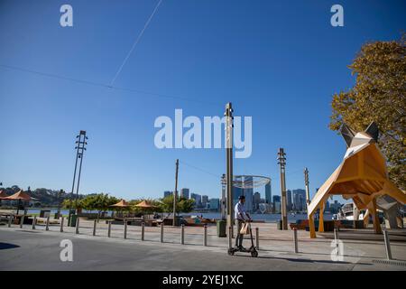 Dieses Foto zeigt einen Mann, der mit einem Elektroroller entlang des Elizabeth Quay Waterfront in Perth fährt, mit einer abstrakten Fuchsskulptur. Stockfoto