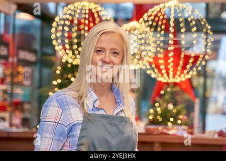 Eine Frau mit blauem Hemd und Schürze steht vor einem Weihnachtsbaum. Sie lächelt und ist glücklich Stockfoto