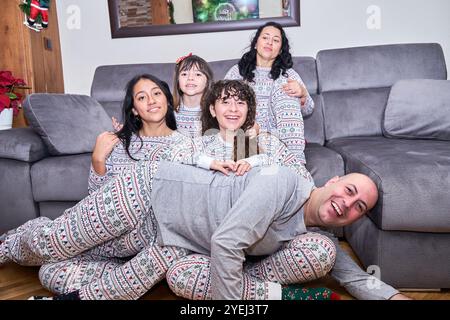 Eine fünfköpfige Familie sitzt im Pyjama auf einer Couch, mit einem Mann auf den Knien und einer Frau auf dem Bauch. Die Szene ist warm und gemütlich, da die Familie Spaß macht Stockfoto