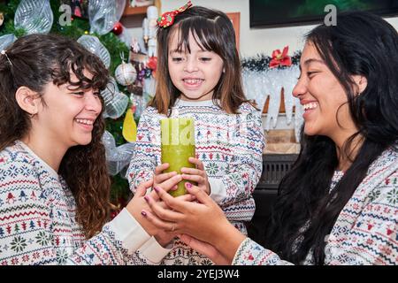 Ein junges Mädchen, das eine grüne Kerze hält, wird von zwei lächelnden Frauen umgeben, die alle festliche Weihnachtspullover vor einem geschmückten Weihnachtsbaum tragen. Stockfoto