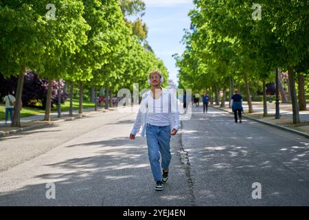 Ein Mann in einem weißen Hemd und einer blauen Jeans läuft die Straße entlang, mit Bäumen im Hintergrund. Er trägt Kopfhörer und hört Musik Stockfoto