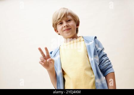 Niedrigwinkelaufnahme eines Jugendlichen Jungen in pastellfarbenen Kleidern, der in die Kamera lächelt, während er ein V-Zeichen vor neutralem Hintergrund im Studio zeigt, im Kopierraum Stockfoto