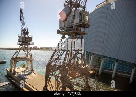 Dieses Foto zeigt zwei alte Industriekräne, die am Ufer positioniert sind, mit dem Wasser und einem Teil eines großen Gebäudes im Hintergrund. Stockfoto