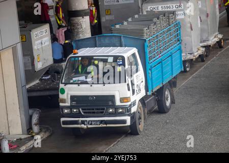 Ein Arbeiter, der einen weißen LKW Daihatsu Delta mit einem blauen Frachtbett in einem Flughafenladebereich betreibt und gestapelte Ausrüstung transportiert. Stockfoto