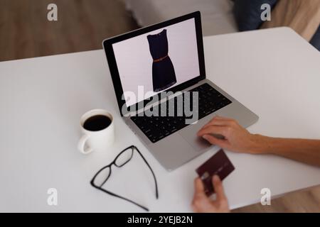 Nahaufnahme von Frauenhänden, die eine Kreditkarte halten. Person, die mit Laptop am Tisch sitzt und beim Online-Shopping ein neues Kleid auswählt. Stockfoto