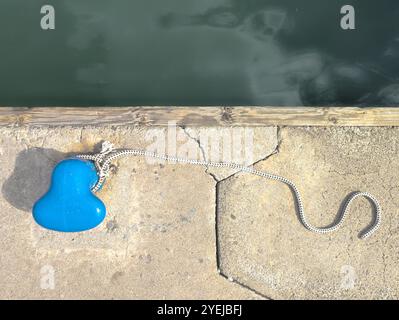 Ein Bild von oben nach unten eines leuchtend blauen Schiffspollers und Seils am Ufer, das zum Binden kleiner Boote verwendet wird, die an einem hellen, sonnigen Tag fotografiert wurden Stockfoto