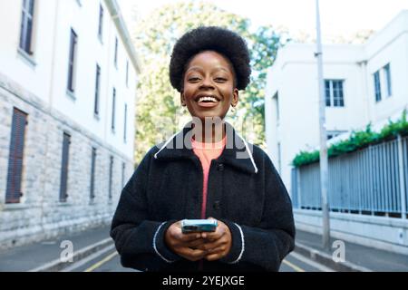 Fröhliche junge Frau lächelt und hält Smartphone in der Urban Street an einem sonnigen Tag Stockfoto