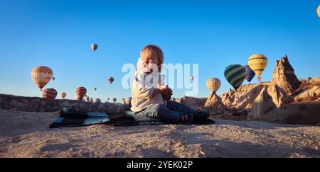 In der wunderschönen Landschaft Kappadokiens spielt ein Kind auf einer Decke, während Dutzende von lebhaften Heißluftballons über den klaren Morgenhimmel gleiten. Stockfoto