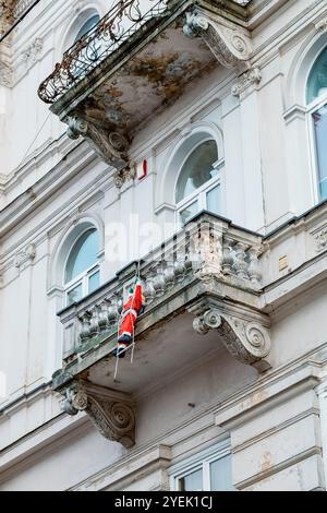Auf dem Balkon eines alten Gebäudes klettert ein Weihnachtsmann auf eine Leiter, weihnachtliche Dekorationen sind sichtbar. Weihnachtliche festliche Einrichtung für die Winterferien Stockfoto