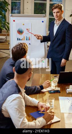 Junger Mann erklärt den verschiedenen Teams globale Marktdaten und stellt anhand eines Flipcharts detaillierte Diagramme und Trends vor Stockfoto