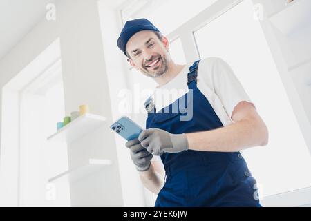 Lächelnder Handwerker in blauen Overalls, der während der Renovierung des Hauses in einem hellen, modernen Apartment ein Smartphone benutzt Stockfoto