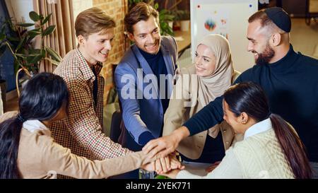 Glückliche, vielfältige Kollegen treffen sich in einem Büro und legen ihre Hände zusammen, um Unterstützung und Verbundenheit zu zeigen. Stockfoto