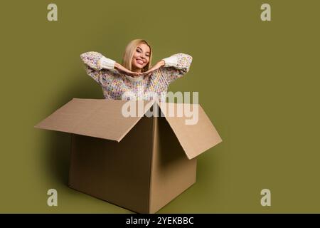 Die lächelnde junge blonde Frau in einem farbenfrohen Strickpullover posiert spielerisch in einem Karton vor khakifarbenem Hintergrund, um Kreativität und Spaß zu genießen Stockfoto