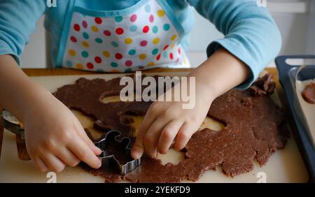 Hände eines 4-jährigen Kaukasiers in einer Schürze, die aus gerolltem Lebkuchenteig verschiedene Formen mit Weihnachtsmetallschneidern schneidet. Der Prozess von Stockfoto