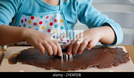 Hände eines 4-jährigen Kaukasiers in einer Schürze, die aus gerolltem Lebkuchenteig verschiedene Formen mit Weihnachtsmetallschneidern schneidet. Der Prozess von Stockfoto