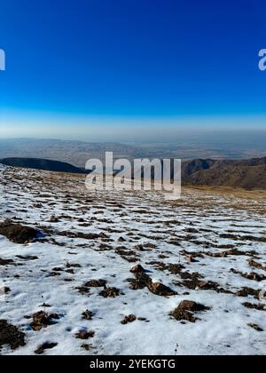 Blick auf Bischkek, bedeckt mit Smog vom Berggipfel, wo sich verschneite Flecken mit felsigem Gelände unter klarem blauen Himmel vermischen Stockfoto