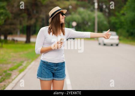 Junge Touristin in einem Sonnenhut innen und Sonnenbrille mit Rucksack, die ein Taxi auf der Straße im Sommer draußen nimmt Stockfoto