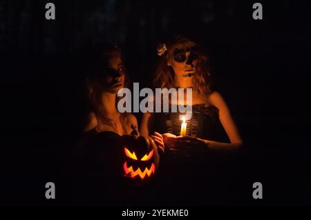 Bizarre zwei-Mädchen-Hexen mit schickem Make-up, die Kürbisse halten, einen gruseligen Jack o' Laterne in einem Wald nachts während Halloween Stockfoto