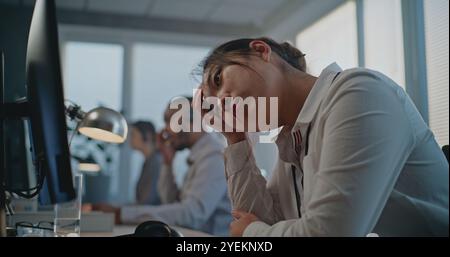 Callcenter-Büro: Dolly-Aufnahme von müdem, gestressten asiatischen Hotline-Operator im Headset mit Computer, beendet den Anruf mit dem Kunden, hält den Kopf. Team aus verschiedenen Kundendienstmitarbeitern. Stockfoto