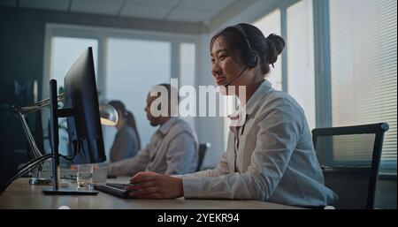 Callcenter-Büro: Dolly-Aufnahme von müdem, gestressten asiatischen Hotline-Operator im Headset mit Computer, beendet den Anruf mit dem Kunden, hält den Kopf. Team aus verschiedenen Kundendienstmitarbeitern. Stockfoto