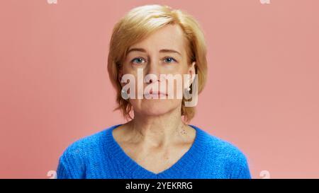Porträt einer reiferen blonden Frau mit blauem Pullover, die mit ernstem und ruhigem Ausdruck vor rosafarbenem Studiohintergrund posiert Stockfoto