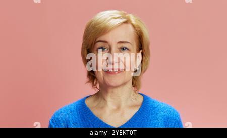 Porträt einer älteren, Reifen Frau mit blonden kurzen Haaren, posiert in blauem Pullover, lächelt vor rosa Studiohintergrund Stockfoto