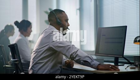 Modernes Callcenter-Büro: Afroamerikanischer Berater setzt sich am Computer, setzt Headset auf, verwendet Computer. Team aus verschiedenen technischen Supportbetreibern, die Online-Hilfe für Kunden anbieten. Stockfoto