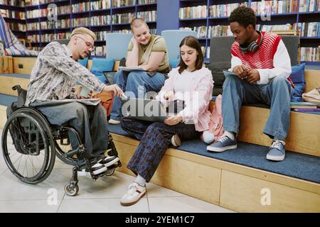 Gruppe verschiedener Studenten, die in einer modernen Bibliothek mit Laptop und Notizbüchern studieren. Teamarbeit und Wissensaustausch auf Holztreppen mit Bücherregalen im Hintergrund Stockfoto