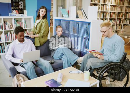 Gruppe von Schülern, die sich mit dem Studium und der Diskussion in der Bibliothek beschäftigen. Multiethnische Gruppen arbeiten zusammen und tauschen Ressourcen aus, wobei ein Rollstuhlfahrer an Lernaktivitäten teilnimmt Stockfoto