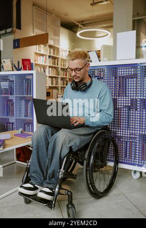 Mann sitzt im modernen Bibliotheksrollstuhl mit Laptop umgeben von Bücherregalen mit Kopfhörern. Konzentriert sich auf Arbeitsaufgaben im öffentlichen Raum Stockfoto