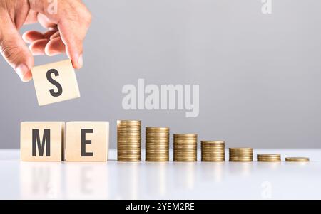 A hand stacking coins with 'SME' written on a wooden block and more coins on the table. Represents financial growth of small and medium-sized enterpri Stockfoto