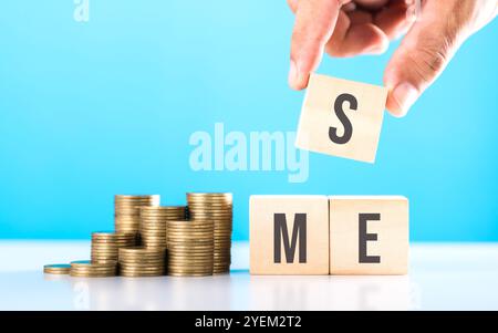A hand stacking coins with 'SME' written on a wooden block and more coins on the table. Represents financial growth of small and medium-sized enterpri Stockfoto