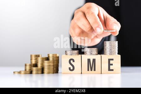A hand stacking coins with 'SME' written on a wooden block and more coins on the table. Represents financial growth of small and medium-sized enterpri Stockfoto