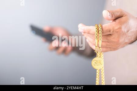 Nahaufnahme eines Mannes, der eine goldene Halskette in der linken Hand hält, während er sein Handy mit der rechten Hand überprüft. Das Konzept der Goldpreisüberwachung, Symbol Stockfoto