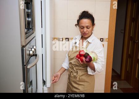 Eine Frau steht in einer Küche und hält frisches Gemüse mit Tomaten und Salat. Sie trägt eine Schürze und scheint bereit zu sein, eine Mahlzeit zuzubereiten. Die Scen Stockfoto