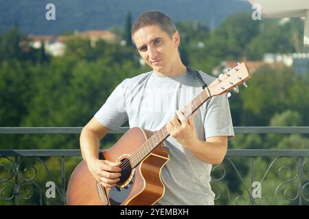 Porträt eines jungen lächelnden Mannes, der draußen an sonnigen Sommertagen steht und Akustikgitarre spielt. Praxis in der Kunst des Musikinstrumentspiels. Stockfoto