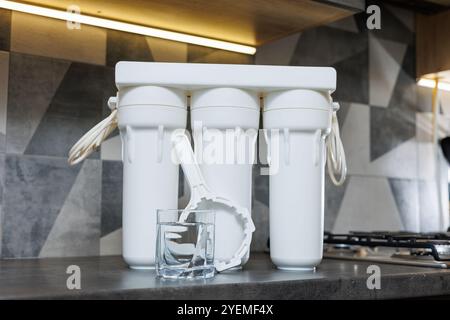 Ein Glas gefiltertes Wasser. Wasserfiltersystem mit austauschbaren Kartuschen. Ein System in der Wohnung mit drei Tanks zur Reinigung von Leitungswasser. Stockfoto