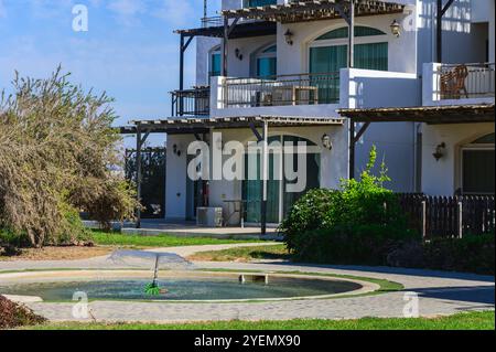 Ein ruhiger Gartenbrunnen ergänzt eine stilvolle Villa, umgeben von üppigem Grün unter der hellen Nachmittagssonne. Stockfoto