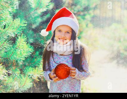 Weihnachten lächelndes Kind, kleines Mädchen mit rotem weihnachtsmann-Hut mit Ball in der Nähe des Baumes Stockfoto