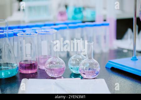 Laborgeräte mit mehreren Glasbehältern, die mit verschiedenfarbigen Flüssigkeiten gefüllt sind. Blaue Reagenzgläser sind im Hintergrund zu anderen Laborgeräten auf dem Tisch sichtbar Stockfoto