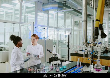 Wissenschaftler in weißen Labormänteln diskutieren experimentelle Ergebnisse in modernen, gut ausgestatteten Labors, umgeben von verschiedenen wissenschaftlichen Instrumenten und Glaswaren Stockfoto