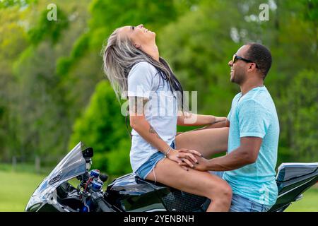 Ein Paar aus mehreren Rassen posiert liebevoll neben ihrem Motorrad auf einem üppigen, grünen Feld. Umarmt, lächeln sie stolz und fangen ihre tiefe Bindung A ein Stockfoto