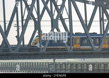 Dieselzug zwischen den Städten, der über Stahlzugbrücke überfährt, Oude IJsselbrug mit trüben niederländischen Landschaften, Fluss IJssel und Auen im hinteren Teil Stockfoto