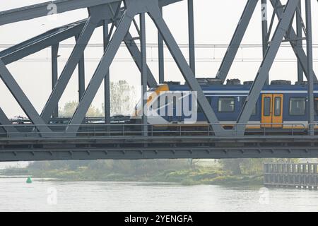 Dieselzug zwischen den Städten, der über Stahlzugbrücke überfährt, Oude IJsselbrug mit trüben niederländischen Landschaften, Fluss IJssel und Auen im hinteren Teil Stockfoto