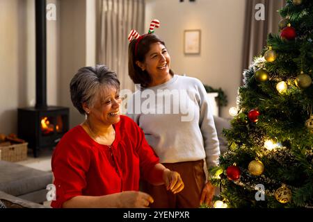 Ältere Freundinnen schmücken den Weihnachtsbaum, lachen zusammen am gemütlichen Kamin, zu Hause. Feiertage, Feier, Freundschaft, fröhlich, glücklich, Stockfoto