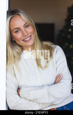 Lächelnde Frau in kuscheliger Pullover, die sich an den Türrahmen in der Nähe des Weihnachtsbaums lehnt. Feiertage, Festtage, Dekoration, Feier, Winter, fröhlich Stockfoto