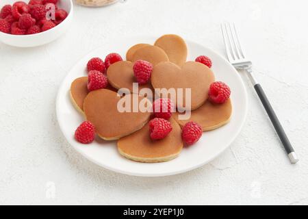 Ein Haufen herzförmiger Pfannkuchen mit Himbeeren und einer Gabel auf dem weißen Teller auf weißem Hintergrund. Valentinstag-Konzept Stockfoto