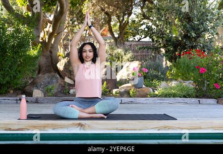 Junge Frau, die Yoga im Freien auf Matte praktiziert, umgeben von üppigem Garten. Wellness, Meditation, Balance, Bewegung, Entspannung, Natur Stockfoto