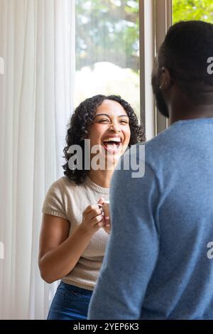 Lachende Frau, die eine Kaffeetasse hält und sich mit dem Partner zu Hause unterhalten kann. Glück, multirassisches Paar, Lebensstil, Bindung, Morgen Stockfoto
