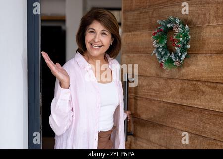 Lächelnde Seniorin begrüßt Gäste zu Hause mit festlichem Weihnachtskranz an der Tür. Feier, Gastfreundschaft, Tradition, Urlaub, fröhlich, Dekoratio Stockfoto